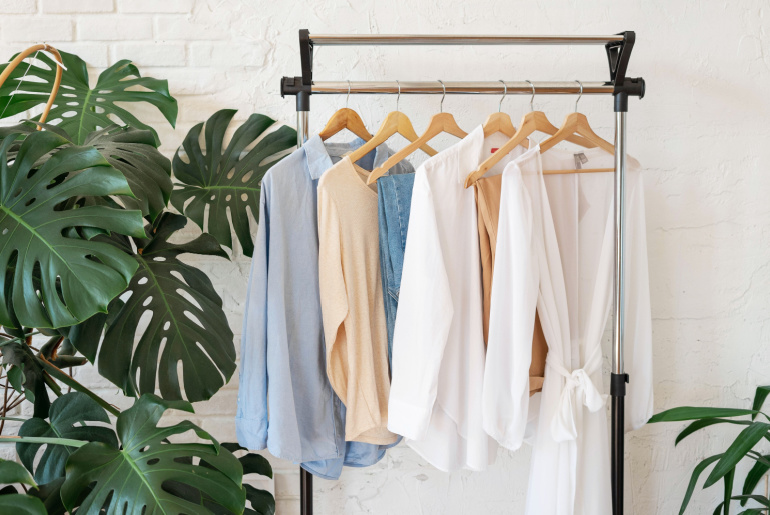 Trendy capsule wardrobe in beige and light blue on a rail rack against the background of a white textured wall with monstera leaves.