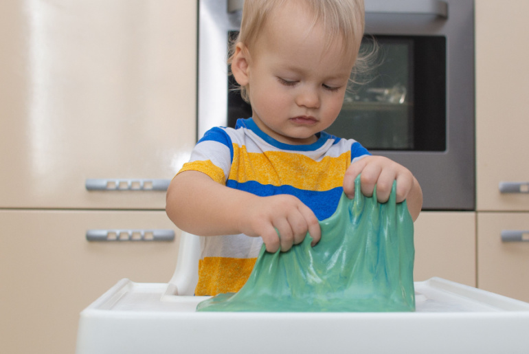 Child play slime at home in the kitchen. Little toddler boy squeeze and stretching green slime sits at a kid's table. Child having fun and being creative plays