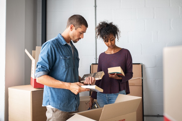 Multiethnic couple reading books while unpacking stuff after relocating to a new flat. Young man and african woman packing cardboard boxes with books. Guy and girl moving house.