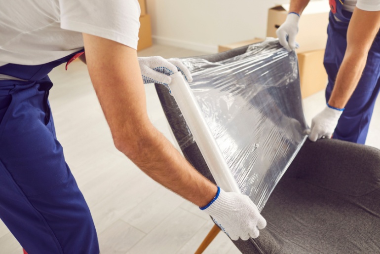 Skilled movers are captured in a close up shot wrapping an armchair in protective stretch film during home or office moving