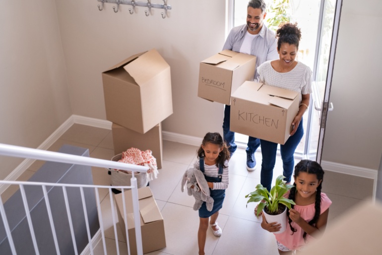Family with two children carrying boxes and plant in new home on moving day. High angle view of happy smiling daughters helping mother and father with cardboard boxes in new house
