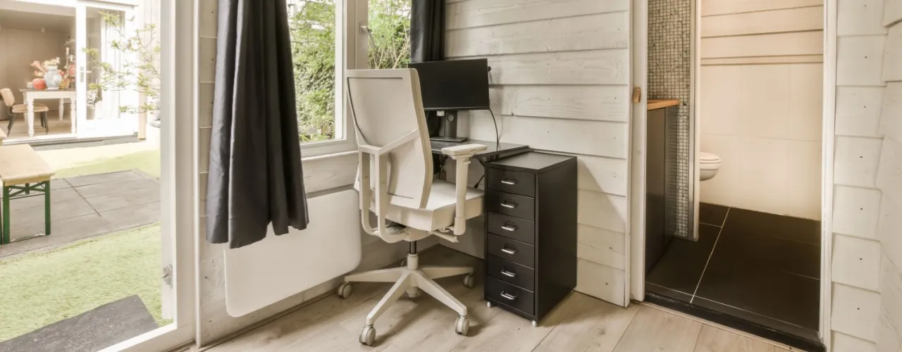 A room with a desk, chair and computer on the floor in front of an open door that leads to a patio.