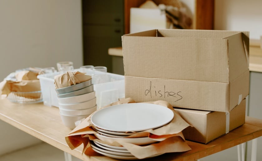 Stacked cardboard boxes labeled dishes and plates, bowls, and glasses on wooden table.