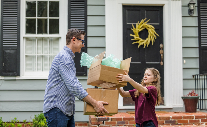 A father and daughter holding moving boxes outside a house.