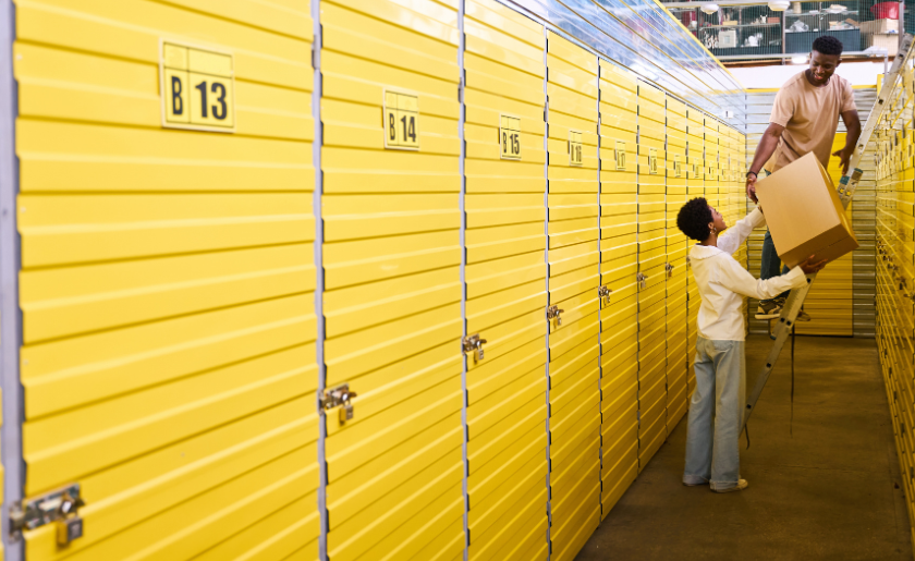 Woman handing box to man on ladder in storage facility with storage lockers.